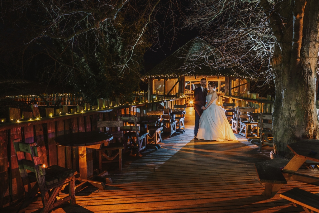 Lights Twinkle at Alnwick Treehouse by night as Married Couple Embrace on the walkway